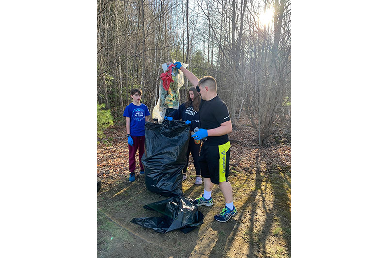 Three students putting garbage into a giant black trash bag outside by some trees